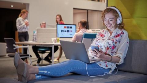 Stock image of a woman smiling while using a computer in an office, with colleagues in the background