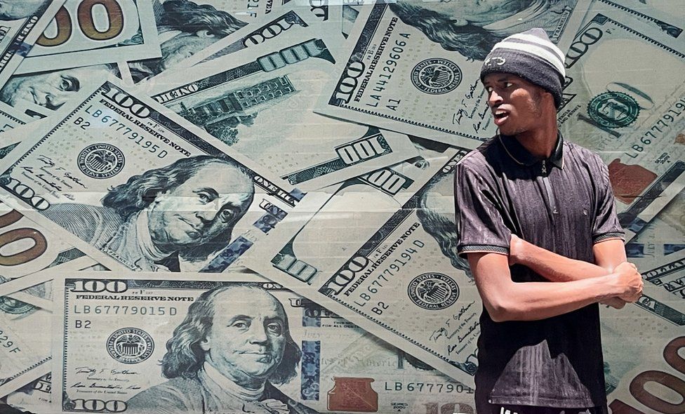 A man stands on the street in front of a currency exchange bureau that is plastered with images of US dollar bills.
