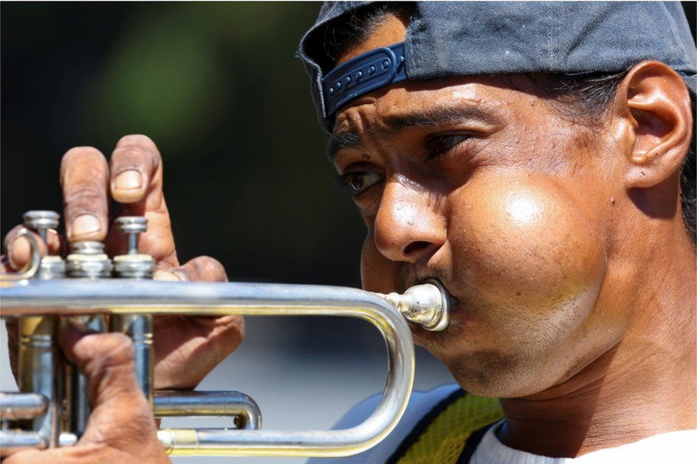 A man plays his trumpet for motorists near traffic lights to earn some money. His cheeks are puffed out as he plays.