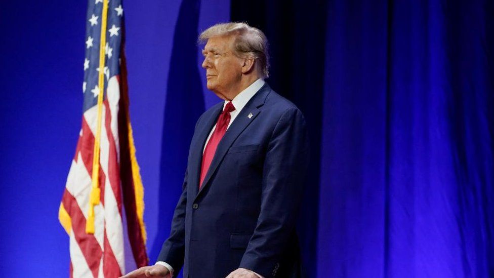 Donald Trump gestures and stands in front of a US flag against a blue backdrop