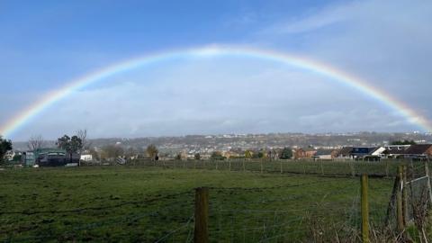 A rainbow arcs across a blue sky with houses below and fields to the front