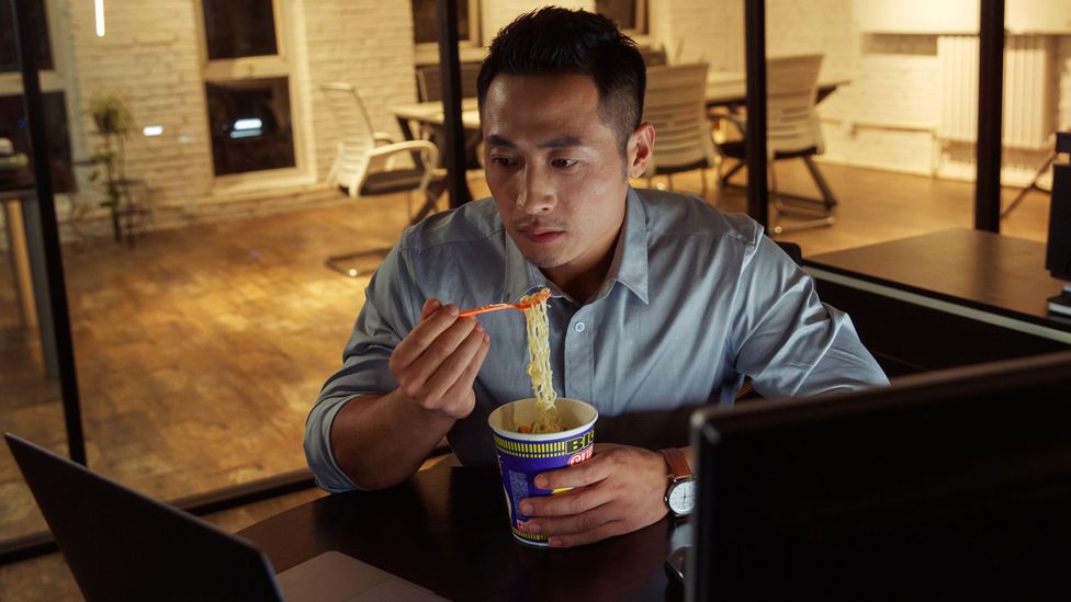 man eats noodle and stares at laptop on desk (Credit: Alamy)